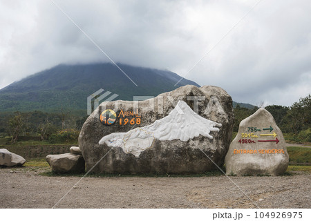Rocks with sign of arenal 1968 trail and directional arrows in front of mountain volcano covered in clouds at Costa Rica Rocks with sign of arenal 1968 trail and directional arrows in front of mountain volcano covered in clouds at Costa Rica 104926975