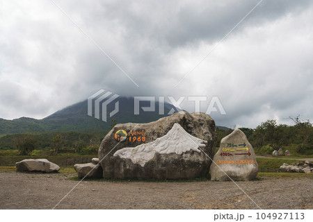 Rocks with sign of arenal 1968 trail and directional arrows in front of mountain volcano covered in clouds at Costa Rica 104927113
