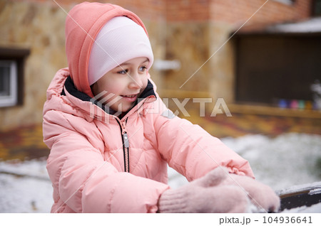 Close-up adorable little kid girl in pink down jacket, makes snowballs while playing outdoor in a snow covered backyard 104936641