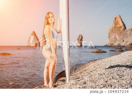 Woman sea sup. Close up portrait of happy young caucasian woman with long hair looking at camera and smiling. Cute woman portrait in a blue bikini posing on sup board in the sea Woman sea sup. Close up portrait of happy young caucasian woman with long hair looking at camera and smiling. Cute woman portrait in a blue bikini posing on sup board in the sea 104937214