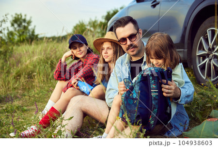 Loving family, man, woman and children sitting by the car on fitness matt and resting after active hiking on hills. Loving family, man, woman and children sitting by the car on fitness matt and resting after active hiking on hills. 104938603