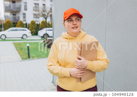 Happy Delivery woman with smartphone and parcel in hands front of the house. Courier send a package to destination. Happy professional shipping courier. Delivery postal service. 104940128