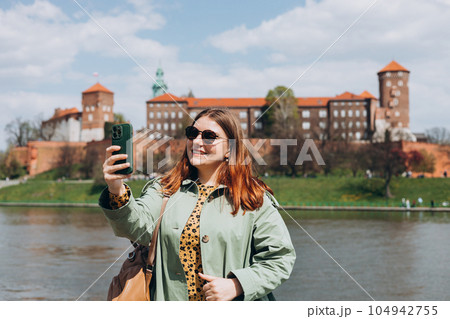 Attractive young female tourist is exploring new city. Redhead girl holding a smartphone in Krakow. Traveling. Happy girl walking in city and makes selfie poses against river and historic building Attractive young female tourist is exploring new city. Redhead girl holding a smartphone in Krakow. Traveling. Happy girl walking in city and makes selfie poses against river and historic building 104942755