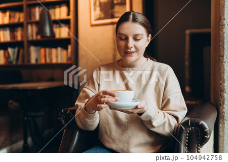 Portrait of joyful young woman enjoying a cup of coffee at home. Smiling pretty girl drinking hot drink in cafe in winter. Excited woman wearing sweater in an autumn day. Portrait of joyful young woman enjoying a cup of coffee at home. Smiling pretty girl drinking hot drink in cafe in winter. Excited woman wearing sweater in an autumn day. 104942758