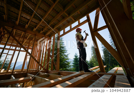 Carpenter constructing wooden frame, two-story house near forest. Bearded man hammering nails with hammer while dressed in protective helmet. Concept of environmentally-friendly modern construction. Carpenter constructing wooden frame, two-story house near forest. Bearded man hammering nails with hammer while dressed in protective helmet. Concept of environmentally-friendly modern construction. 104942887