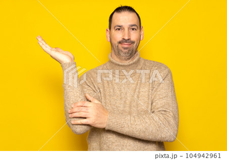 Handsome latino man offering something imaginary in the palm of his hand with friendly and affable attitude, isolated on yellow studio background Handsome latino man offering something imaginary in the palm of his hand with friendly and affable attitude, isolated on yellow studio background 104942961