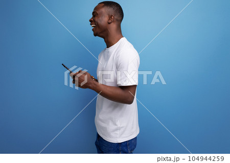 casual 30s african man with short haircut in white t-shirt chatting while holding smartphone in hand casual 30s african man with short haircut in white t-shirt chatting while holding smartphone in hand 104944259