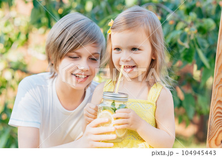 Children drink natural lemonade at stand in park. Summer refreshing natural drink lemonade. Detox fruit infused flavored water, cocktail in a beverage dispenser with fresh fruits Children drink natural lemonade at stand in park. Summer refreshing natural drink lemonade. Detox fruit infused flavored water, cocktail in a beverage dispenser with fresh fruits 104945443