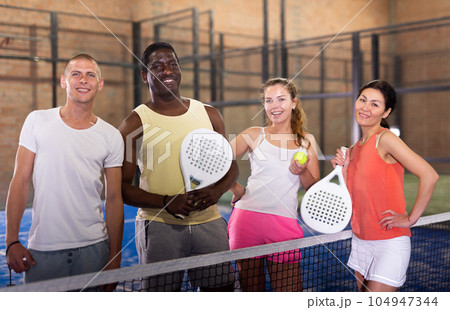 Smiling men and women with rackets and balls posing on indoor padel court Smiling men and women with rackets and balls posing on indoor padel court 104947344