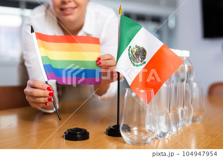 A woman in a white blouse with friendly smile arrangs flags of Mexico and LGBT for meeting. A woman in a white blouse with friendly smile arrangs flags of Mexico and LGBT for meeting. 104947954