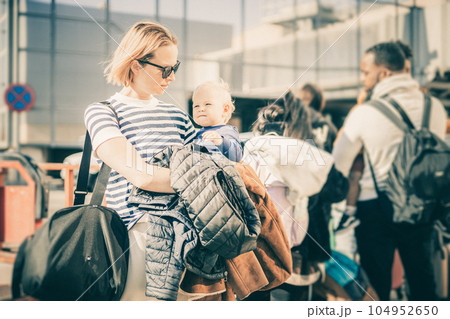 Motherat travelling with his infant baby boy child. Mom holding travel bag and her infant baby boy child while queuing for bus in front of airport terminal station Motherat travelling with his infant baby boy child. Mom holding travel bag and her infant baby boy child while queuing for bus in front of airport terminal station 104952650