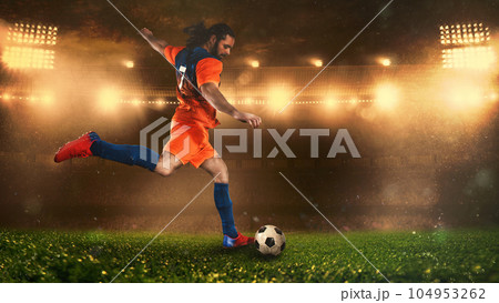 Soccer scene at night match with player in a orange uniform kicking the ball with power 104953262