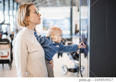 Mother traveling with child, holding his infant baby boy at airport terminal, checking flight schedule, waiting to board a plane. Travel with kids concept Mother traveling with child, holding his infant baby boy at airport terminal, checking flight schedule, waiting to board a plane. Travel with kids concept 104954417