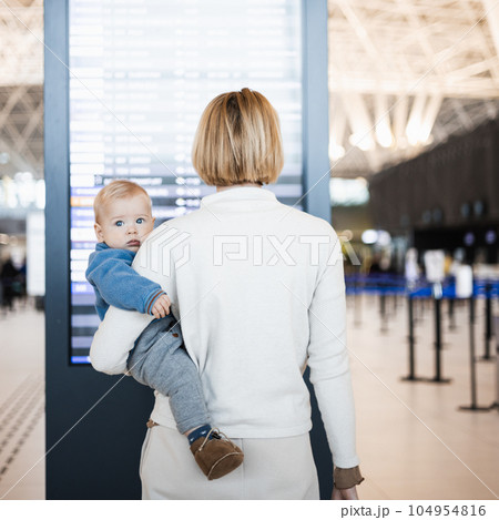 Mother traveling with child, holding his infant baby boy at airport terminal, checking flight schedule, waiting to board a plane. Travel with kids concept 104954816
