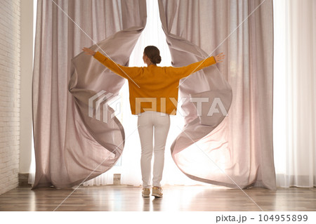 Woman opening window curtains at home in morning, back view 104955899