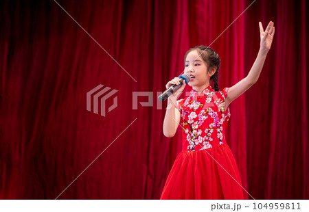 The Asian kid girl sing a song on stage at her school activity day, dress in Qipao style, red curtain background The Asian kid girl sing a song on stage at her school activity day, dress in Qipao style, red curtain background 104959811