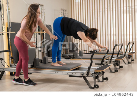 Overweight caucasian woman doing pilates exercises on reformer with personal trainer.  104962445