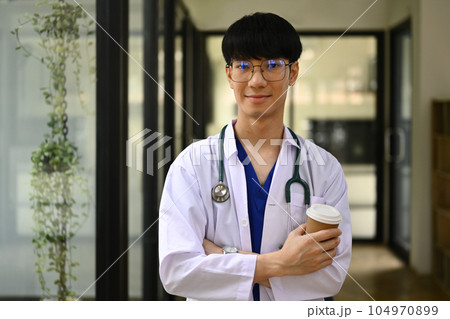 Smiling general practitioner in white coat with stethoscope standing at hospital hallway and smiling to camera 104970899