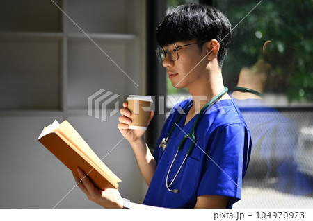 Medical student man wearing blue scrubs reading book, preparing for university exams. Medical internship concept Medical student man wearing blue scrubs reading book, preparing for university exams. Medical internship concept 104970923