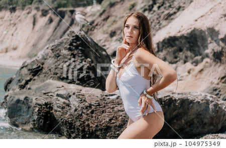 Woman travel sea. Happy tourist in white bikini enjoy taking picture outdoors for memories. Woman traveler posing on the beach at sea surrounded by volcanic mountains, sharing travel adventure journey 104975349