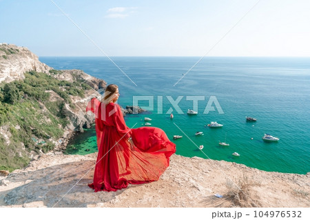 Red Dress Woman sea Cliff. A beautiful woman in a red dress and white swimsuit poses on a cliff overlooking the sea on a sunny day. Boats and yachts dot the background. Red Dress Woman sea Cliff. A beautiful woman in a red dress and white swimsuit poses on a cliff overlooking the sea on a sunny day. Boats and yachts dot the background. 104976532