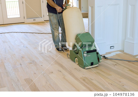 Carpenter grinding wooden parquet floor by using floor sander an newly constructed house Carpenter grinding wooden parquet floor by using floor sander an newly constructed house 104978136