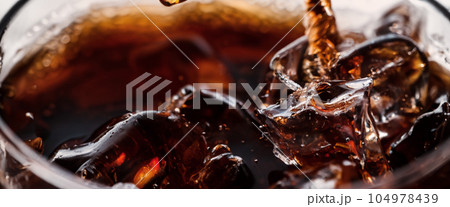 Pouring of Cola and Ice. Cola soda and ice splashing fizzing or floating up to top of surface. Close up of ice in cola water. Texture of carbonate drink with bubbles in glass. Cold drink background 104978439