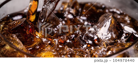 Pouring of Cola and Ice. Cola soda and ice splashing fizzing or floating up to top of surface. Close up of ice in cola water. Texture of carbonate drink with bubbles in glass. Cold drink background Pouring of Cola and Ice. Cola soda and ice splashing fizzing or floating up to top of surface. Close up of ice in cola water. Texture of carbonate drink with bubbles in glass. Cold drink background 104978440