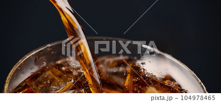 Pouring of Cola and Ice. Cola soda and ice splashing fizzing or floating up to top of surface. Close up of ice in cola water. Texture of carbonate drink with bubbles in glass. Cold drink background Pouring of Cola and Ice. Cola soda and ice splashing fizzing or floating up to top of surface. Close up of ice in cola water. Texture of carbonate drink with bubbles in glass. Cold drink background 104978465