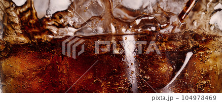 Pouring of Cola and Ice. Cola soda and ice splashing fizzing or floating up to top of surface. Close up of ice in cola water. Texture of carbonate drink with bubbles in glass. Cold drink background Pouring of Cola and Ice. Cola soda and ice splashing fizzing or floating up to top of surface. Close up of ice in cola water. Texture of carbonate drink with bubbles in glass. Cold drink background 104978469