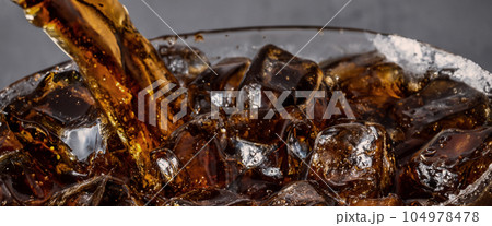 Pouring of Cola and Ice. Cola soda and ice splashing fizzing or floating up to top of surface. Close up of ice in cola water. Texture of carbonate drink with bubbles in glass. Cold drink background Pouring of Cola and Ice. Cola soda and ice splashing fizzing or floating up to top of surface. Close up of ice in cola water. Texture of carbonate drink with bubbles in glass. Cold drink background 104978478