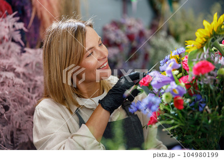 Smiling woman flower shop owner in apron looking on bouquet of flowers at florist store Smiling woman flower shop owner in apron looking on bouquet of flowers at florist store 104980199