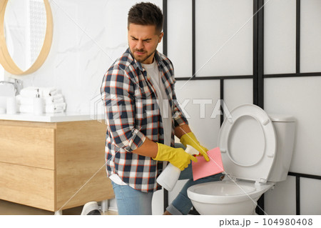 Young man feeling disgust while cleaning toilet bowl in bathroom Young man feeling disgust while cleaning toilet bowl in bathroom 104980408