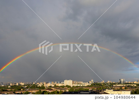 A large semicircle of a rainbow in the gray sky above the roofs of city houses. 104980644
