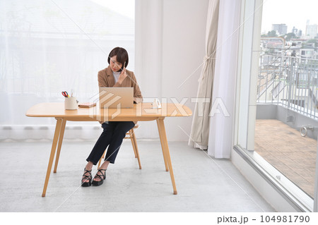屋内の木製の机でノートパソコンとスマホで電話をする女性 屋内の木製の机でノートパソコンとスマホで電話をする女性 104981790