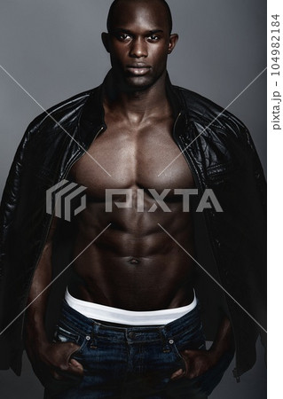 Sexy, fashion model and portrait of a black man in a jacket isolated on a dark background in studio. Serious, handsome and an African person modeling leather clothes and jeans with confidence 104982184