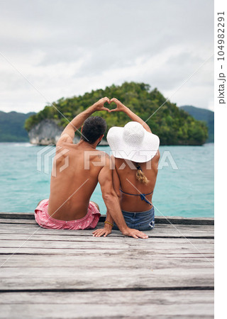Heart shape, hands and couple by ocean while on a summer, romantic and tropical vacation. Island, dock and back of man and woman relaxing on wood pier with love gesture on holiday or weekend trip. Heart shape, hands and couple by ocean while on a summer, romantic and tropical vacation. Island, dock and back of man and woman relaxing on wood pier with love gesture on holiday or weekend trip. 104982291