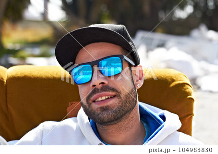 Man, portrait and sunglasses with smile on street for urban fashion or cool summer style in the city. Happy male person or face of stylish model with cap smiling and relaxing on chair in the outdoors 104983830