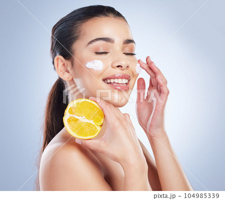 Lemon, skincare and asian woman with face cream in studio for wellness or cosmetic mask on grey background. Citrus, fruit and lady with facial for dark spots, melasma or acne prevention or sunscreen 104985339
