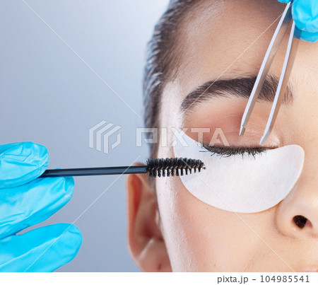 Woman, lashes and tweezer with mascara, studio closeup and makeup artist in gloves, hands and extension. Girl, model and zoom of face for beauty, cosmetics and brush for aesthetic by gray background 104985541