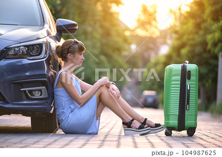 Young tired woman with suitcase sitting near her car waiting for someone. Travel and vacations concept. 104987625