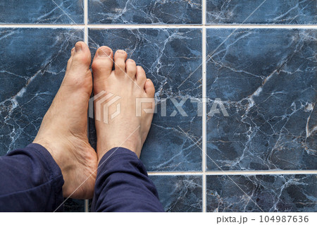 Young man crossing feet on square tiles. Veins in feet visible. 104987636