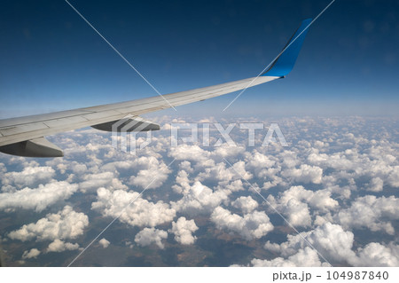 View of jet airplane wing from inside flying over white puffy clouds in blue sky. Travel and air transportation concept. 104987840