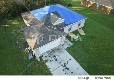 Top view of leaking house roof covered with protective tarp sheets against rain water leaks until replacement of asphalt shingles. Damage of building rooftop as aftermath of hurricane Ian in Florida Top view of leaking house roof covered with protective tarp sheets against rain water leaks until replacement of asphalt shingles. Damage of building rooftop as aftermath of hurricane Ian in Florida 104987960