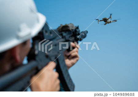 Caucasian man in a helmet shoots a flying drone with a rifle. Caucasian man in a helmet shoots a flying drone with a rifle. 104989688