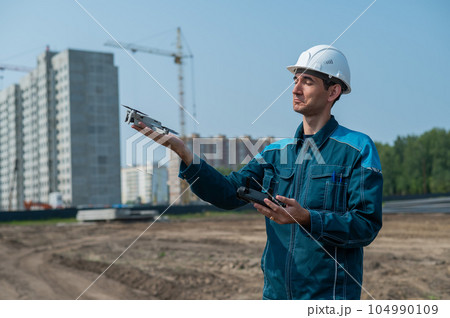 A man in a helmet and overalls controls a drone at a construction site. The builder carries out technical oversight. 104990109