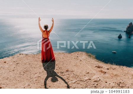 Woman in red dress on sea. Side view a Young beautiful sensual woman in a red long dress posing on a rock high above the sea on sunset. Girl on the nature on blue sky background. Fashion photo. 104991183