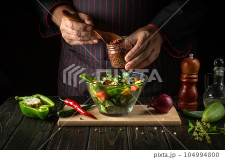 The cook prepares vegetarian dishes from fresh vegetables. Menu concept for a hotel on a dark background. Adding dry aromatic spices to salad. The cook prepares vegetarian dishes from fresh vegetables. Menu concept for a hotel on a dark background. Adding dry aromatic spices to salad. 104994800