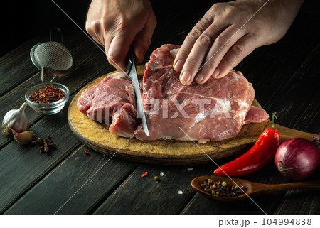 The cook cuts raw meat steak with a knife on the wooden cutting board of the restaurant kitchen. The concept of the process of preparing a meat dish for lunch with aromatic spices and pepper. The cook cuts raw meat steak with a knife on the wooden cutting board of the restaurant kitchen. The concept of the process of preparing a meat dish for lunch with aromatic spices and pepper. 104994838