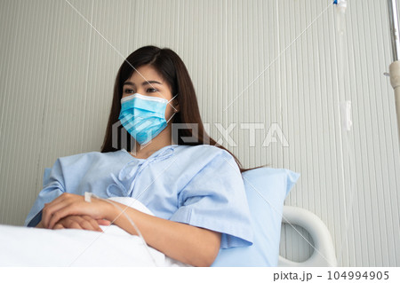 Sad Asian patient woman lying on the hospital bed and wearing a face mask to protect coronavirus. Concept of Health care, quarantine coronavirus (COVID-19), and Health insurance. 104994905